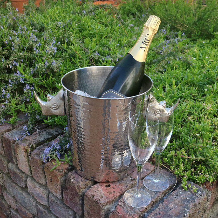Champagne bottle in a rhino-themed ice bucket with two flutes, set on a brick wall with greenery.