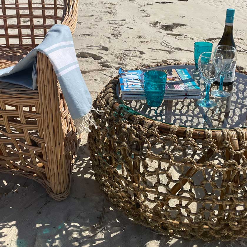 Wicker chair and table on sandy beach with wine, glasses, and magazines.