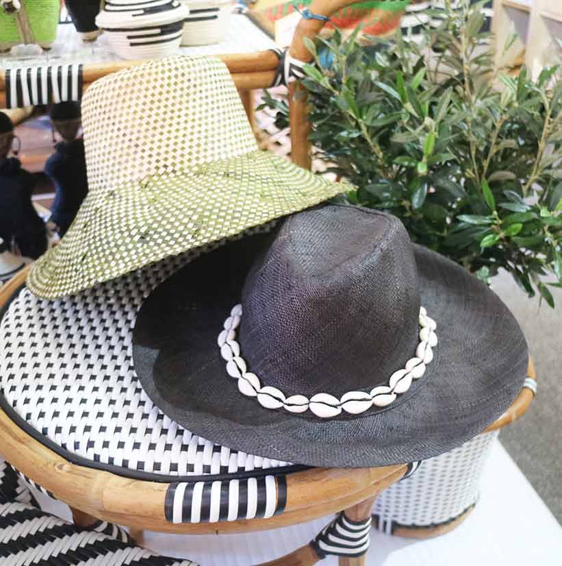 Two stylish hats on a woven chair, one green and white, the other black with shell trim, surrounded by greenery.